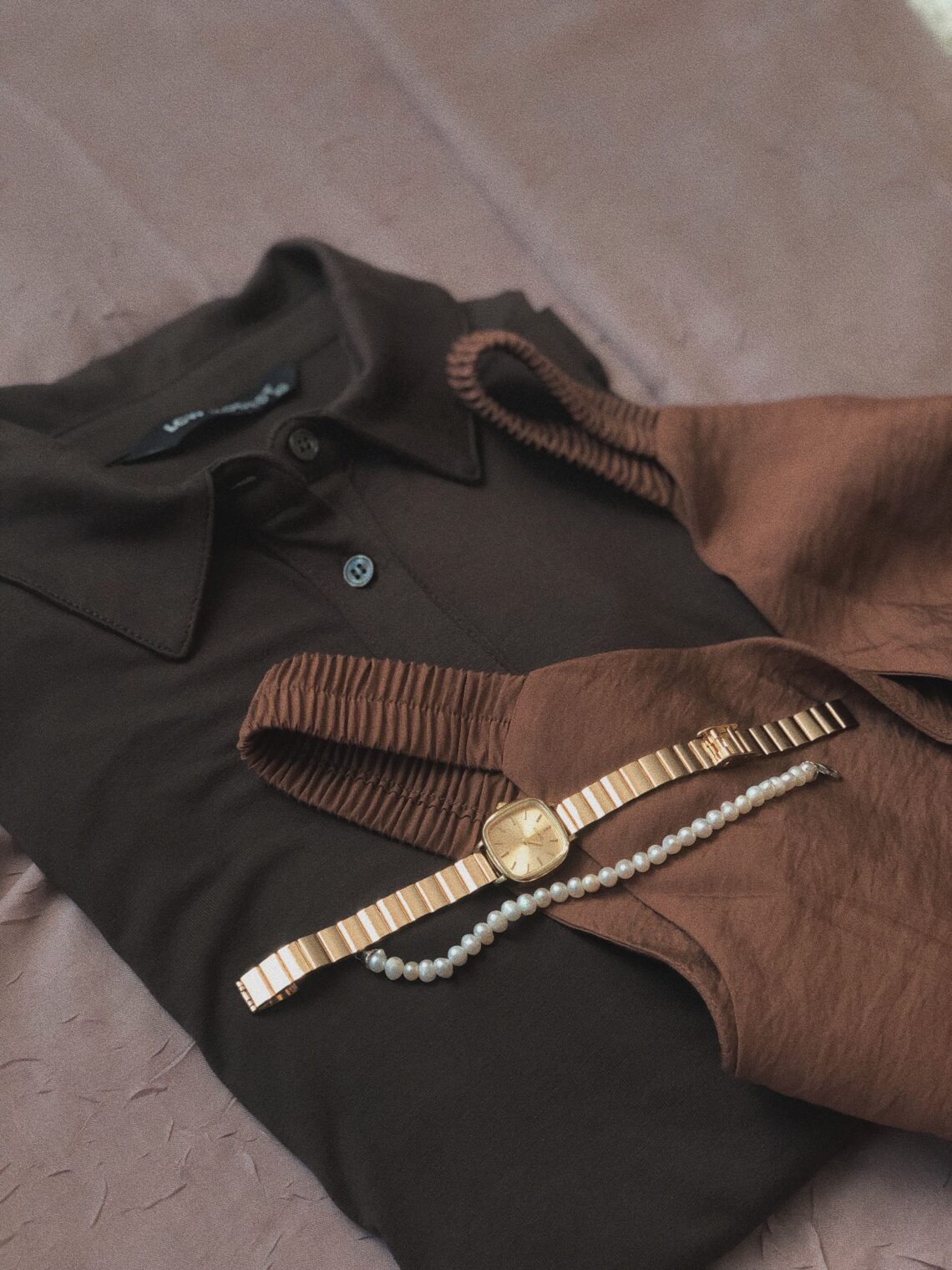 Warm neutral flat lay of a gold watch and pearl necklace draped over a soft brown knit sweater